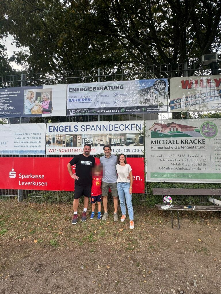 Drei Erwachsene und ein Kind im Trikot vor der Sponsorenbande mit dem Visato-Banner